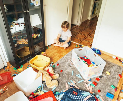 Young child sitting in a cluttered playroom, looking bored despite being surrounded by too many toys